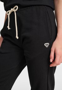 Schwarze Jogginghosen aus strukturiertem Material mit einem Kordelzugbund und cremefarbenen Bändern sowie einem kleinen gestickten Logo an der Hüfte.