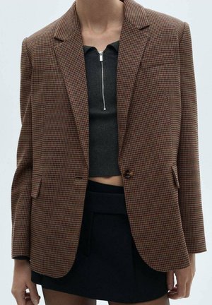 Person wearing a brown checkered blazer over a black cropped top with a front zipper and a black skirt against a plain background.