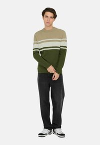 Gebreide trui met beige, groene en witte horizontale strepen, ronde halslijn en lange mouwen; gecombineerd met donkergrijze jeans en sneakers.