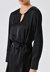 Abito in satin nero con scollo a chiave, maniche lunghe e una cintura legata in vita. Presenta un accento decorativo in metallo dorato.