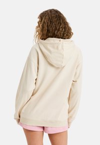 Beige hoodie met een relaxte pasvorm, met lange mouwen en geribde manchetten, gedragen over roze shorts. Zacht materiaal met een gladde textuur.