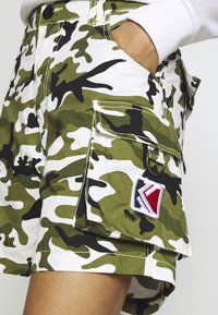 Pantaloni cargo mimetici verdi e bianchi realizzati in tessuto, con accenti neri, molteplici tasche e una colorata toppa con logo ricamato.
