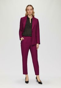Un costume sur mesure magenta avec un blazer à un bouton, un pantalon tapered et un blouse plissée noire. Des escarpins noirs brillants complètent le look.