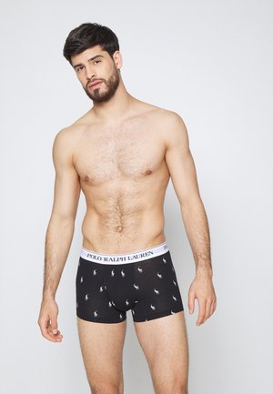 Homme torse nu avec des cheveux bruns et une barbe portant un slip noir Polo Ralph Lauren avec une ceinture en logo blanche et de petites impressions blanches.