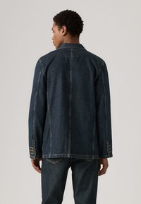 Σκοτεινό τζιν μπουφάν με ευθεία γραμμή, κουμπιά στους καρπούς και ορατά ραφές. Διαθέτει γιακά και είναι στιλιζαρισμένο με τζιν.