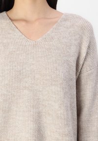 Femme portant un pull en maille côtelée beige avec un col en V, montrant le décolleté et le haut de la poitrine sur un fond blanc uni.