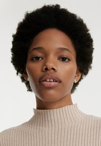 Beige geribbelde top met hoge hals, gestructureerde stof, gedragen met kleine gouden oorbellen. Natuurlijke haarkleur met korte krullen, neutrale huidtint, zachte uitdrukking.