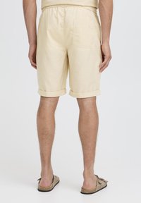 Pantaloncini di lino giallo chiaro con risvolti e tasca applicata sul retro, indossati con sandali marrone chiaro. Presentano una cintura elastica.