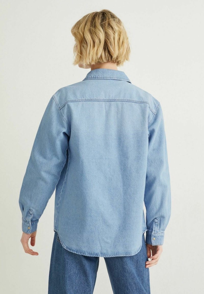 Personne aux cheveux blonds courts portant une chemise en denim bleu clair et un jean bleu, vue de dos sur un fond uni.