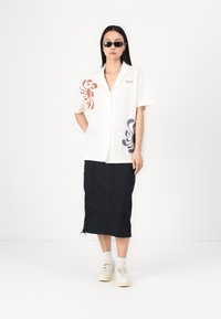 Blouse crème à manches courtes avec imprimé floral rouge et bleu marine, associée à une jupe noire jusqu'aux genoux avec poches latérales et détail à cordon.