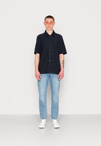 Camisa preta de botão de manga curta, jeans azul claro e ténis brancos. Os acessórios incluem óculos de sol redondos e com lentes rosa.