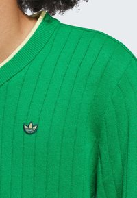 Grøn ribbet sweater med V-hals og en kontrasterende kremfarvet krave. Har et broderet logo i hvidt og sort på brystet.