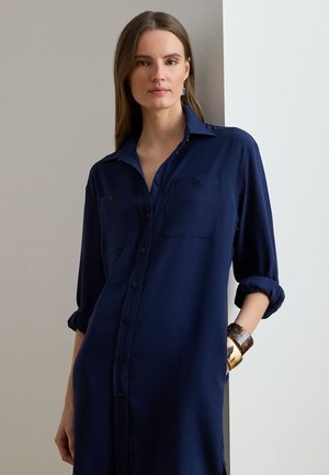 Granatowa koszulowa sukienka z długim rękawem z miękkiego materiału z guzikami, dwoma kieszeniami na piersiach i podwiniętymi rękawami. Uzupełniona brązową i złotą bransoletką na mankiecie.