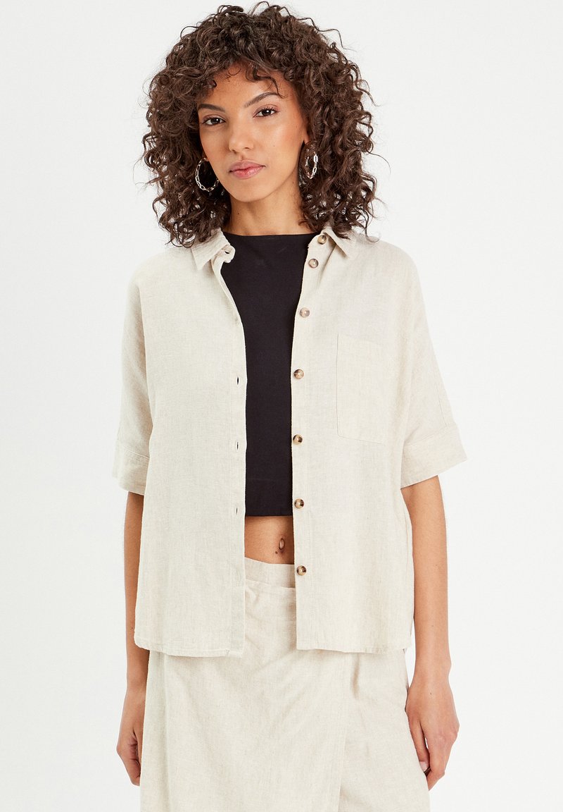Chemise en lin à boutons, couleur blanc cassé, manches courtes, poche poitrine simple, portée avec un crop top noir, assortie à un bas beige.