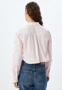 Camisa corta de color rosa claro con mangas largas, que presenta lazos de tela en los brazos, puños abotonados y un pliegue en la parte posterior. Llevada con pantalones de mezclilla azules.