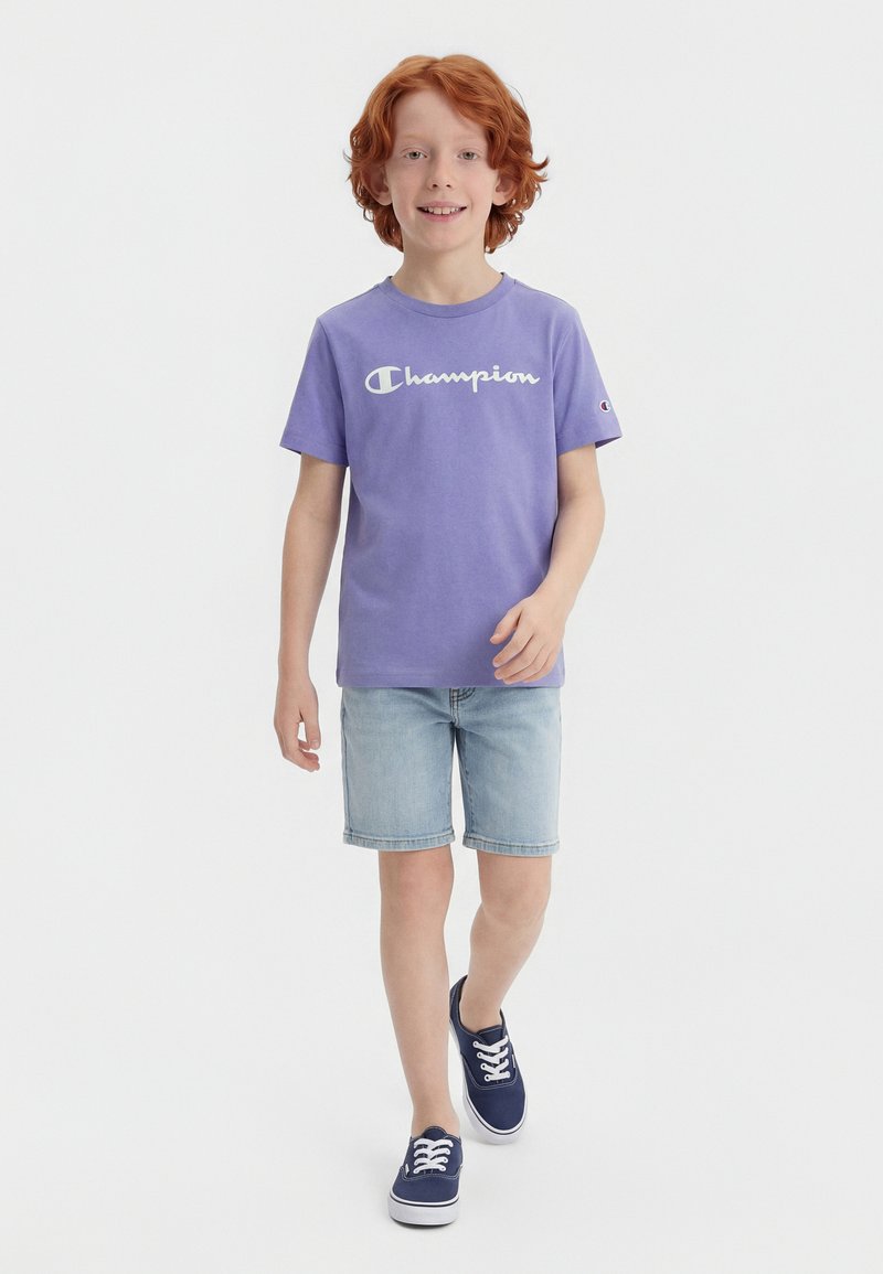 Garçon aux cheveux roux marchant, portant un t-shirt violet Champion, un short en denim clair et des baskets bleu marine sur un fond blanc.