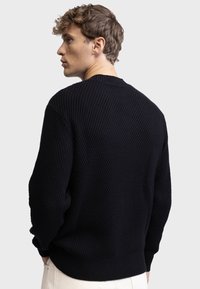 Pull en maille noir avec un motif texturé, encolure ras du cou et poignets et ourlet côtelés. Porté à l'envers, montrant les détails à l'arrière.