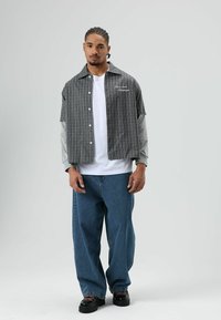 Kariertes Kurzarmhemd mit grauen Ärmeln, weißem T-Shirt mit Grafik darunter, lockere Jeans und schwarze klobige Schuhe.