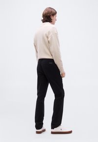 Άνδρας με καστανά μαλλιά μεσαίου μήκους στέκεται στραμμένος προς τα αριστερά, φορώντας ένα μπεζ πουλόβερ, μαύρο παντελόνι και λευκά αθλητικά παπούτσια με καφέ σόλες.