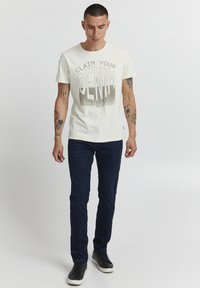 T-shirt crème avec un grand imprimé graphique portant le texte "RECLAMEZ VOTRE DENIM" en gris foncé, associé à un jean slim bleu marine et des baskets noires.