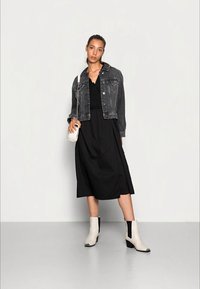 Vestido preto midi com uma jaqueta de ganga, combinado com botas de tornozelo. A jaqueta tem um corte clássico; o vestido é pregueado na cintura. A bolsa branca realça o visual.