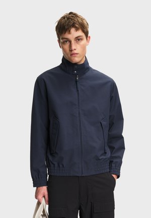 Ung mand iført en marineblå lynlåstrøje med høj krave og elastiske ærmekanter, der holder en beige taske i venstre hånd mod en ensfarvet baggrund.