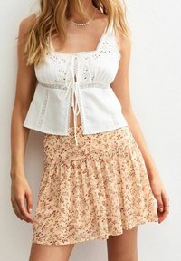 Žena v bílé bezrukávové topu s krajkovým vázáním a krémové sukně s vysokým pasem s růžovým květinovým potiskem a volánkem na lemu.