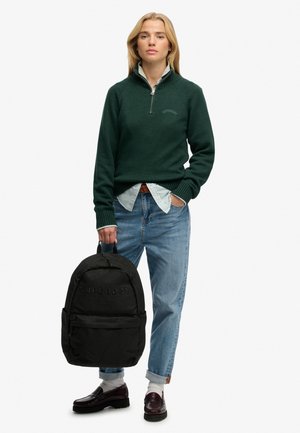 Donkergroene trui met ritskraag, lichte blauw gestreepte blouse eronder, gecombineerd met een blauwe spijkerbroek en een zwarte rugzak.