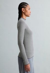 Vrouw met gevlochten haar, gekleed in een langarmig, geribd zwart-wit gestreept shirt en grijze broek, staand in profiel tegen een effen achtergrond.