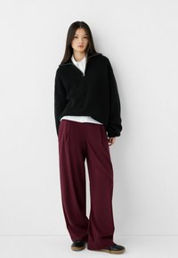Pull en tricot noir avec un col zippé, associé à un pantalon bordeaux ample et des chaussures noires avec une semelle texturée.