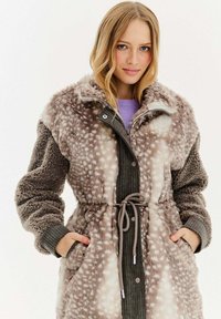 Mladá žena s blond vlasy, oblečená v hnědém a bílém puntíkovaném kabátu z umělé kožešiny, s tkaničkovým pasem a rukama v kapsách.