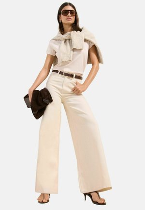 Kvinde iført beige bukser med vide ben, lyse t-shirt, striktrøje over skuldrene, brun bælte og hæle, holder en mørkebrun clutch, solbriller på.