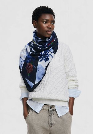 Bufanda triangular de suave tela con un patrón floral azul y un borde oscuro. Lleva puesta alrededor del cuello sobre un suéter blanco de punto cable.