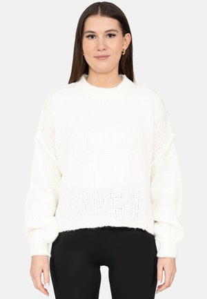 Jonge vrouw met lang bruin haar, gekleed in een witte gebreide trui en een zwarte broek, staand tegen een egale lichte achtergrond.