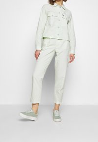 Light green, textured jacket with two chest pockets, paired with matching light green pants and gray-green sneakers with white soles.