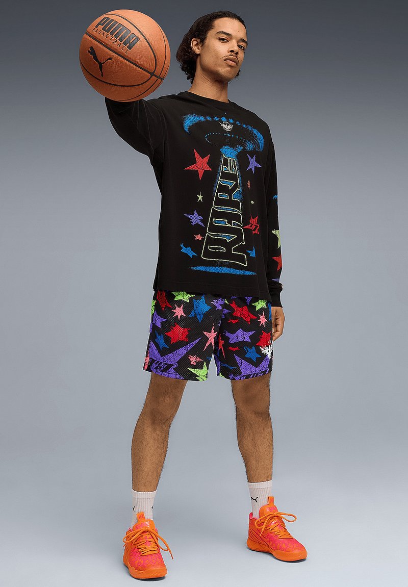 Schwarzes Langarmshirt mit bunten Stern-Grafiken und dem Text "RARE". Shorts mit mehrfarbigem Sternenmuster und leuchtend orangefarbenen Turnschuhen. Hält einen Basketball.