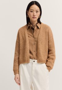 Hellbrauner gestrickter Cardigan mit lässiger Passform, offenem Vorderteil und geripptem Saum. Über einem hellbeigen gestreiften Hemd getragen, kombiniert mit weißen Hosen.
