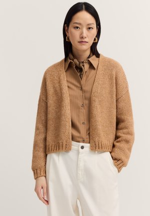 Hellbrauner gestrickter Cardigan mit lässiger Passform, offenem Vorderteil und geripptem Saum. Über einem hellbeigen gestreiften Hemd getragen, kombiniert mit weißen Hosen.