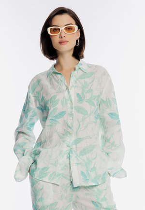 Kobieta ubrana w białą lnianą koszulę i spodnie z nadrukiem jasnych zielonych liści, pomarańczowe prostokątne okulary przeciwsłoneczne oraz srebrne kolczyki koła, ręce w kieszeniach.