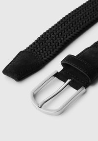 Melnā austā josta ar teksturētu dizainu, zamšādas galiem un sudraba metāla sprādzi ar diviem izvirzītiem asmeņiem. Acīmredzama šuvju detaļa.