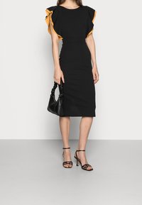 Musta tiukkalaatuinen mekko, jossa on keltaiset röyhelöhiat, yhdistettynä mustaan käsilaukkuun ja nauhakenkiin. Sileä pinta; elegantti siluetti.