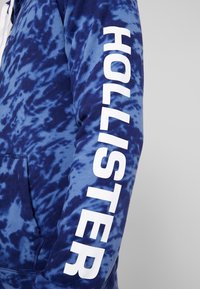 Haut à capuche bleu marine tie-dye avec une poche avant ; présente le texte blanc "HOLLISTER" le long de la manche gauche. Texture de tissu douce.