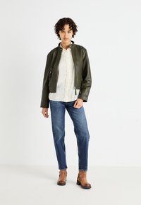 Chaqueta bomber de cuero verde oliva, blusa blanca con volantes, jeans rectos azules y botines marrones de tacón con detalles de correa.