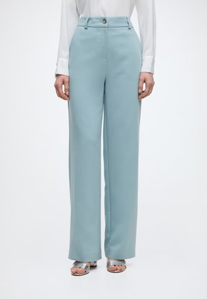 Femme portant un pantalon bleu clair taille haute à jambes larges, un chemisier blanc à manches longues et des talons ouverts argentés, debout sur fond blanc.