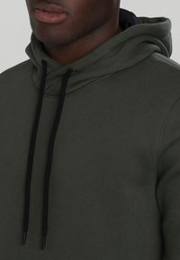 Sudadera de color verde oscuro con una textura suave, que presenta cordones negros y un bolsillo tipo canguro. Diseño sin costuras con ningún logo o patrón visible.