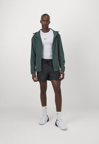 Jachetă verde cu glugă, tricou alb sportiv cu un logo, şorturi negre cu model, adidaşi albi şi şosete albe. Îmbrăcăminte sport simplă şi funcţională.