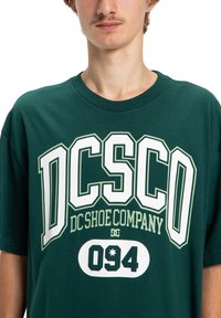 Ciemnozielona bawełniana koszulka z wyraźnym białym i jasn zielonym tekstem: "DC Shoe Company" oraz "094", z okrągłym logo. Klasyczny krój z okrągłym dekoltem.