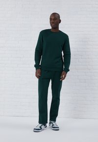 Mørkegrøn sweatshirt og matchende bukser, lavet af et blødt materiale. Har ribbede manchetter og bund, med hvide sneakers der står i kontrast til outfittet.