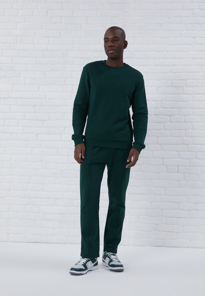 Felpa verde scuro e pantaloni coordinati, realizzati in un materiale morbido. Presentano polsini e orlo a costine, con sneakers bianche che contrastano l'outfit.