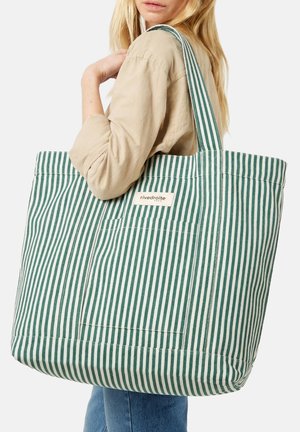 Femme blonde en chemise beige tenant un grand sac cabas rayé vertical vert et blanc avec poche avant et étiquette sur l'épaule.